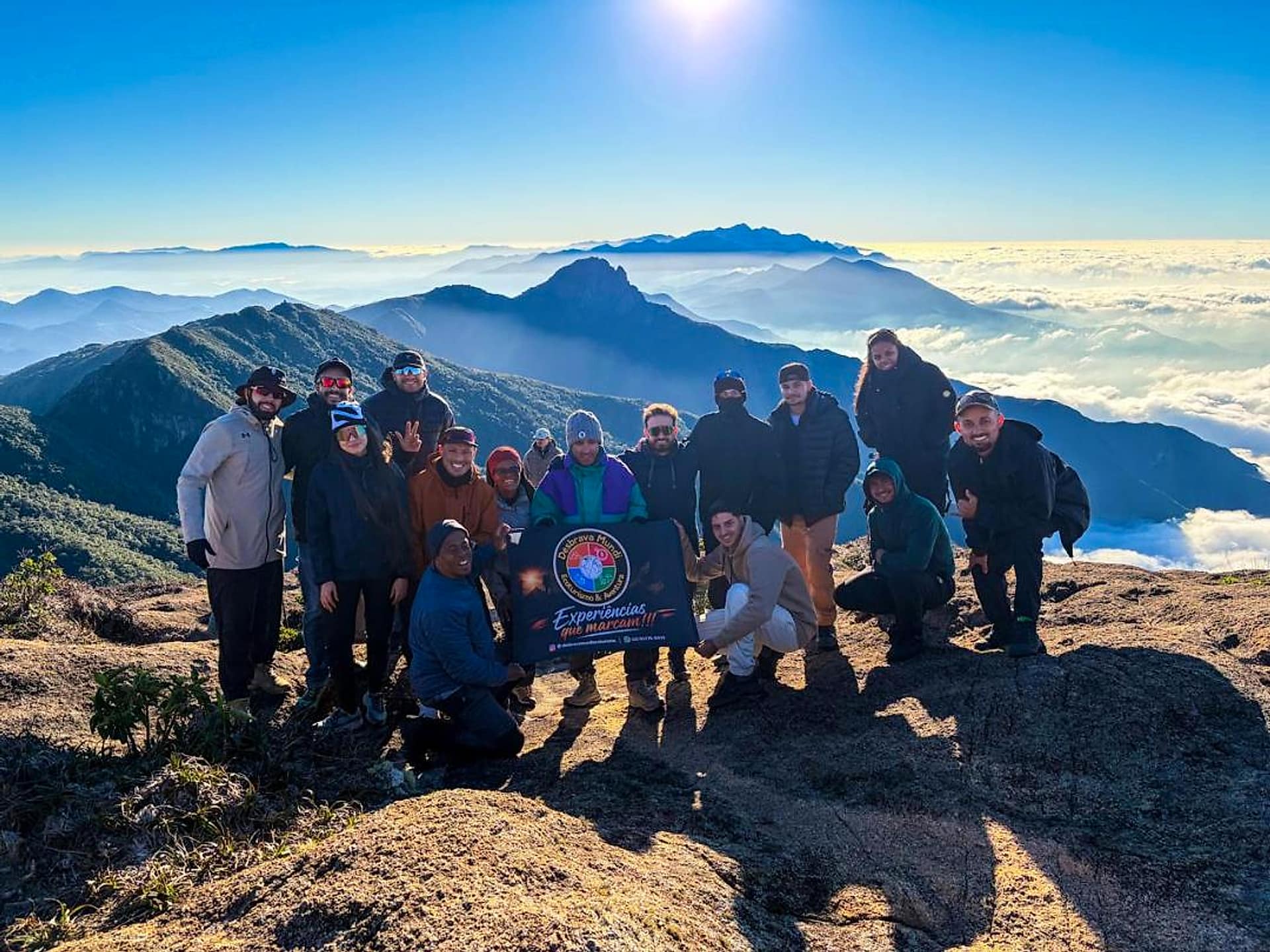 Turma da Desbrava Mundi com a faixa da agência no cume, acima de um mar de nuvens
